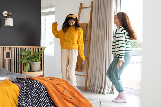 Two female friends wearing mustard sweaters playing in bedroom near bed with orange blanket