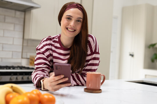 Woman wearing maroon striped top leaning on marble kitchen island, holding mauve smartphone, mug