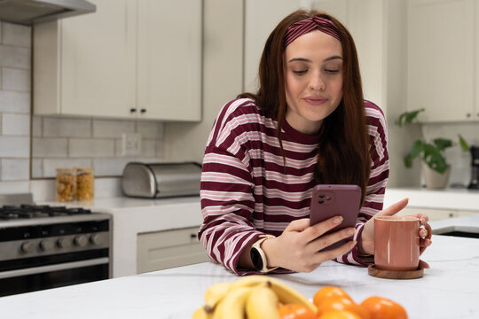 Adult female wearing striped top leaning in home kitchen, holding mauve phone and pink mug, fruit