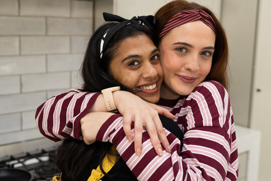 Diverse female friends hugging wearing maroon-striped tops and headbands in kitchen by gas stove