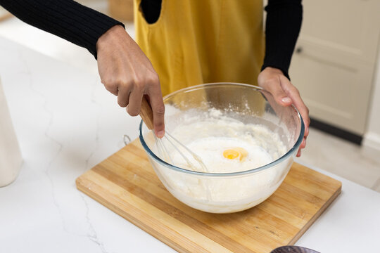 Female in yellow apron whisking batter in glass bowl on cutting board in kitchen, cracked egg