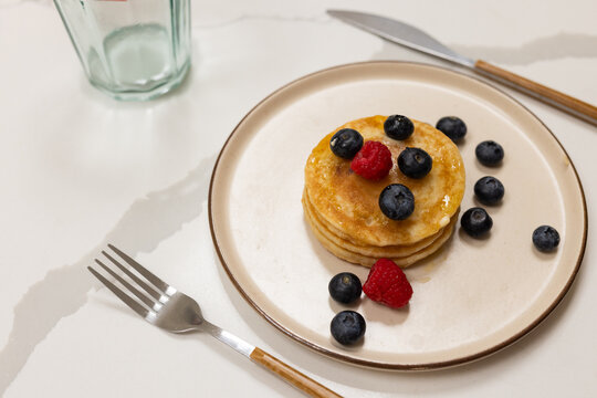 Four pancake stack is sitting on pale countertop near fork and knife, syrup and berries