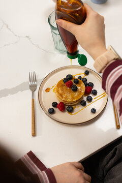Woman pouring syrup from squeeze-bottle onto pancakes with berries on marble top, striped top watch