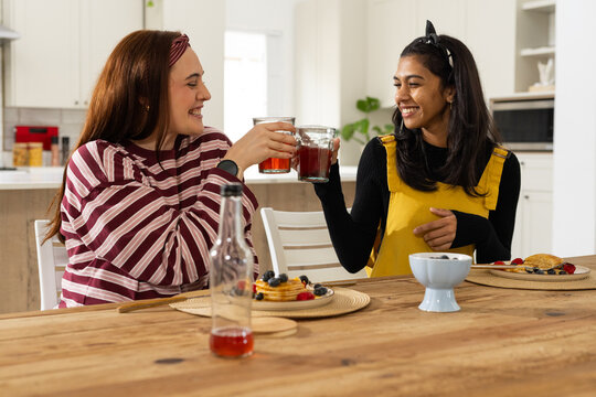 Diverse female friends clinking glasses, sitting at dining table in bright kitchen with pancakes
