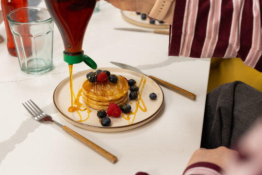 Adult woman pouring amber syrup from green bottle onto pancakes on white table striped sleeve watch