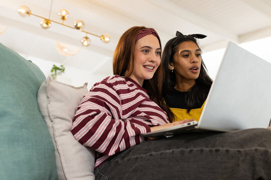 Female friends sitting on couch sharing laptop at home, wearing striped top and yellow overalls