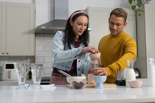 Couple wearing denim jacket and mustard sweater making smoothie at kitchen island with blender jar