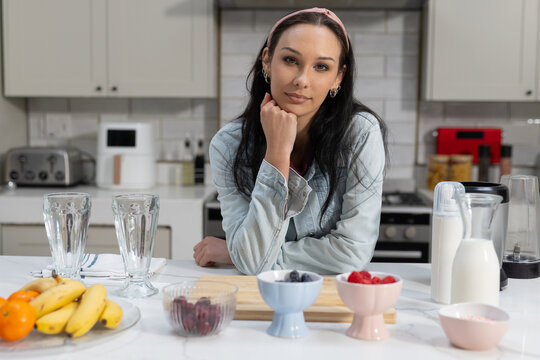 Adult woman leaning on kitchen island, resting chin, presenting smoothie prep bowls and blender