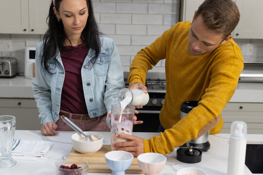 Couple in yellow sweater and denim jacket pouring milk into blender cup with strawberry on counter