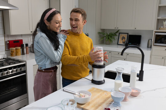 Couple operating personal blender at kitchen island, man in mustard sweater, woman in denim jacket