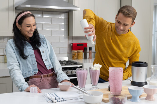 Couple making pink smoothies at home kitchen man dispensing whipped cream can while woman watching