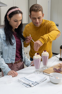 Couple preparing pink milkshakes in parfait glasses at kitchen countertop, adding cherries