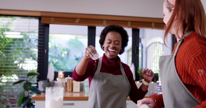 Diverse female friends sprinkling from shaker, rolling dough with pin at counter making cookies