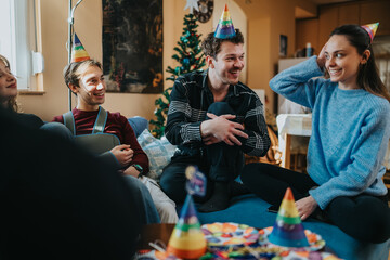 Naklejka premium A group of friends wearing party hats laugh and chat during a home birthday celebration. The cozy living room scene shows relaxed conversation and festive decorations around a Christmas tree.