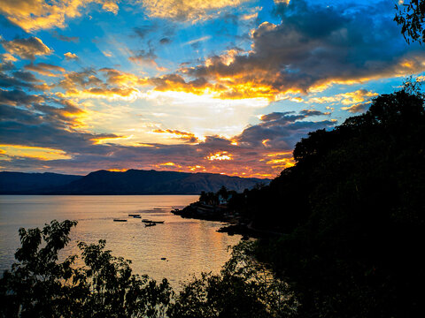 Golden sun dips low over calm waters. Clouds glow in amber, blue, and deep purple hues. Silhouetted trees frame the shoreline gently. Distant hills stand quiet beneath the fading light
