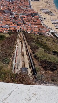 Two Nazar&eacute; funicular cars cross on the hillside, with one ascending while the other simultaneously travels in the opposite, descending direction toward the town and beach in Portugal.