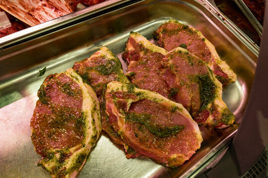 Several raw pork steaks are placed in a metal tray, marinated with herbs and spices. The meat appears fresh and seasoned with a green herb mixture, ready for cooking or grilling. The close-up view hig