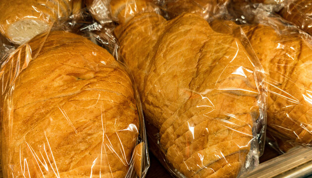 Several loaves of sliced bread wrapped in clear plastic packaging, displayed together. The bread has a golden brown crust and a soft texture, indicating freshness. Grouping of different types of bread