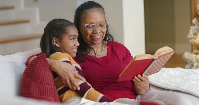Senior African American woman and teen granddaughter reading aloud on sofa holding red book bonding