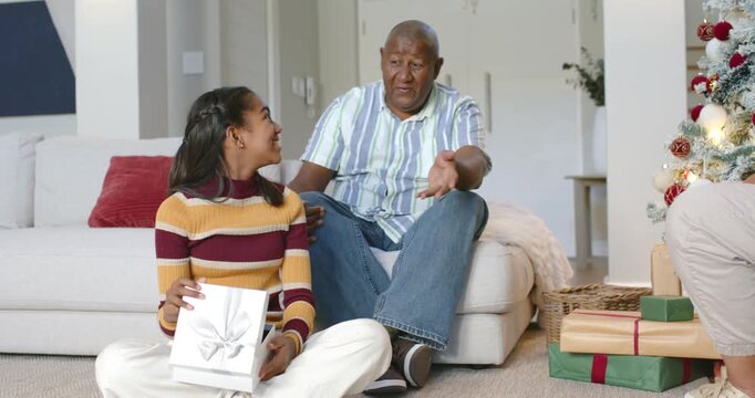 African American senior man sitting at home, gesturing and urging teen girl opening holiday gift