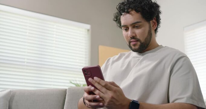African American man late twenties holding red phone, tapping and leaning on sofa reading article