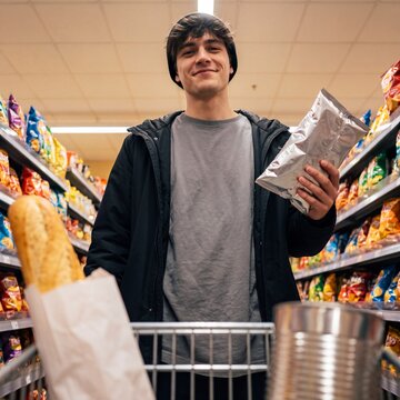 A young man in a beanie and jacket smiles as he shops in a grocery store, holding a bag of chips and with a loaf of bread in his shopping cart amidst colorful snack aisles.