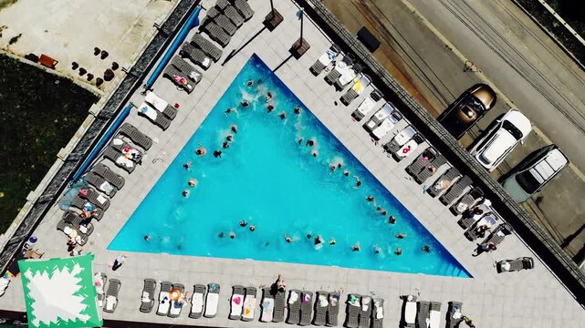 People relax by a large triangular pool in a sunny location with sunbeds and umbrellas