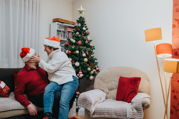 Naklejka premium A middle-aged couple wearing Santa hats share a tender, joyful moment on a couch beside a decorated Christmas tree. They laugh and embrace in a cozy living room decorated for the holidays.