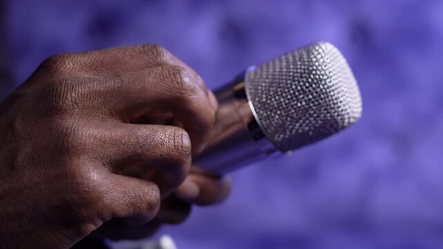 Close-up of Hand Holding Microphone - A person's hand, with visible skin texture, holds a silver microphone against a blurred purple background.