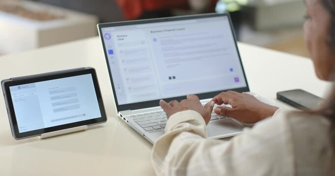 Hands are reaching and tapping laptop keyboard at desk, are typing to respond to tablet chat