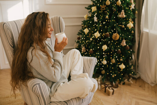 Young beautiful woman drinking tea sitting under decorated Christmas tree.