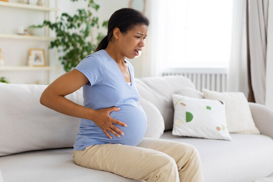 Pregnant Black woman sits on a sofa at home with painful expression, holding her belly and lower back while experiencing physical discomfort during pregnancy