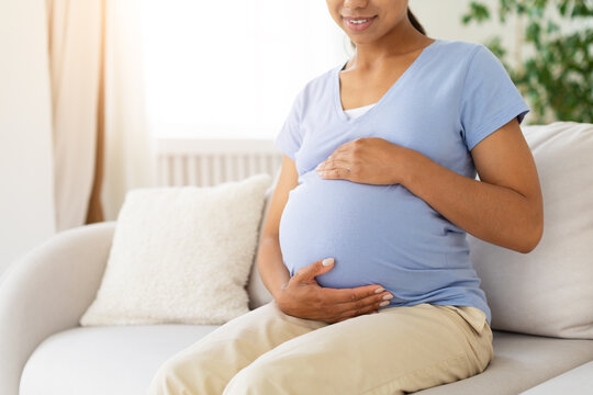 Close-up of pregnant Black woman sitting on sofa at home, gently cradling her baby bump with both hands while relaxing in a bright and comfortable living room