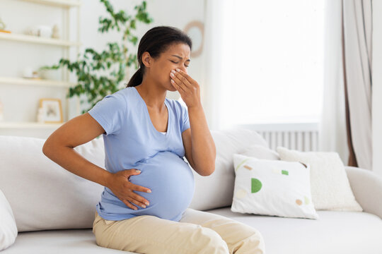 Pregnant Black woman sitting on sofa at home, covering her mouth and holding her belly while appearing nauseous and uncomfortable during a difficult moment in pregnancy