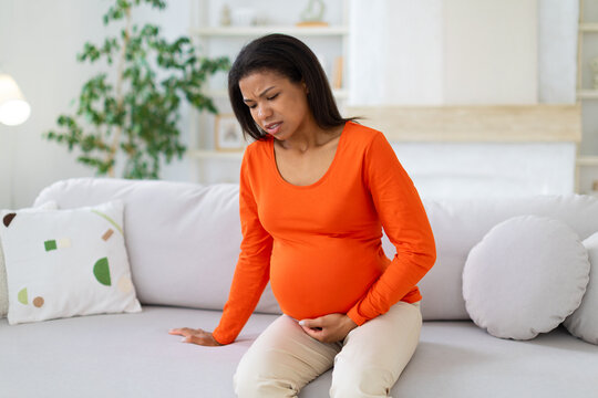 Pregnant Black woman sitting on sofa at home with painful expression, supporting her lower abdomen while experiencing discomfort during a difficult moment in pregnancy