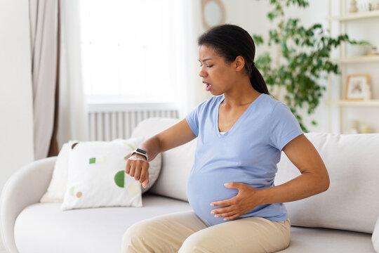 Pregnant woman sitting on sofa at home checking smartwatch, timing contractions while holding belly with concerned expression