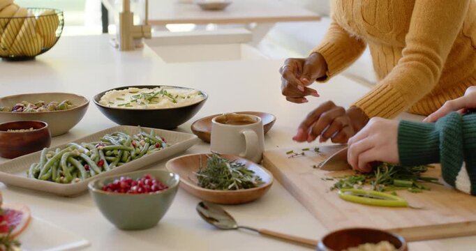 Diverse friends prompted by bowls chopping herbs with chef knife at counter prepping garnishes