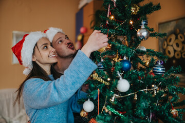 Naklejka premium A young couple decorates a Christmas tree together at home, both wearing Santa hats and smiling. They hang ornaments and lights on the festive tree in a cozy holiday interior.