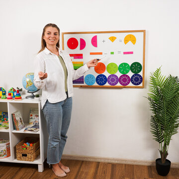 Female teacher explains fractions using colorful fraction circles and strips arranged on a whiteboard, demonstrating visual mathematics and arithmetic during a classroom lesson.
