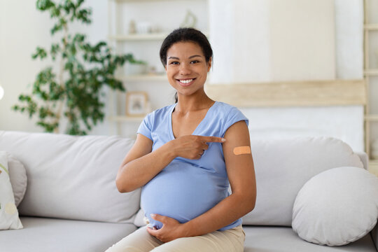 Smiling pregnant Black woman sits on a sofa at home, pointing to a bandage on her upper arm after vaccination while holding her baby bump during prenatal healthcare