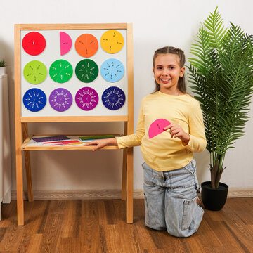 Smiling schoolgirl holds a one-half fraction piece beside a board with colorful fraction circles, demonstrating hands-on visual mathematics learning and fraction concepts.