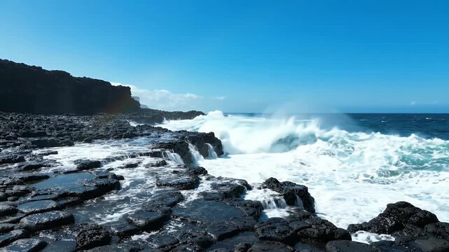 Crashing Waves on Rocky Shoreline Under Blue Sky - Powerful ocean waves crash against a dark, hexagonal rock formation on a coastal shoreline.