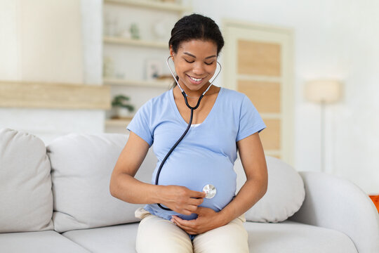 Smiling pregnant Black woman sitting on sofa at home, using stethoscope on her baby bump to listen to the fetal heartbeat during a gentle prenatal self-check