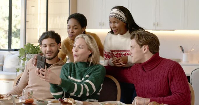 Diverse friends at kitchen table watching woman in green knit taking selfie showing phone laughing