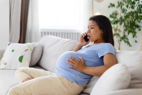 Pregnant Black woman speaking on smartphone with surprised expression while holding her baby bump at home during a concerning maternity moment