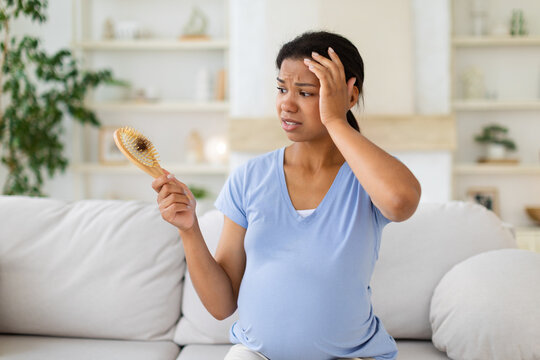 Worried pregnant Black woman sits on a sofa at home, examining hair collected in brush and touching her head while concerned about hair loss during pregnancy