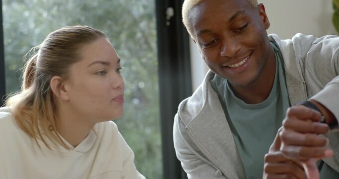 Diverse couple sitting near glass door, man examining smartwatch, woman reaching for smartphone