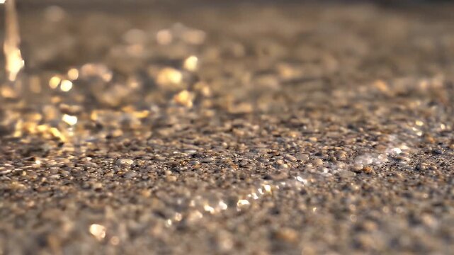 Close-up Of Gentle Waves Washing Over Tiny Shells On Sandy Beach - Shallow ocean waves with shimmering sunlight gently recede from a beach covered in small shells and pebbles.
