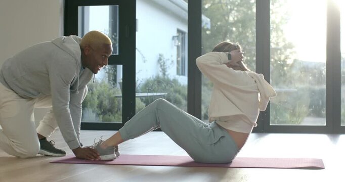 African American woman doing crunches on pink mat with coach holding shoes coaching by glass doors