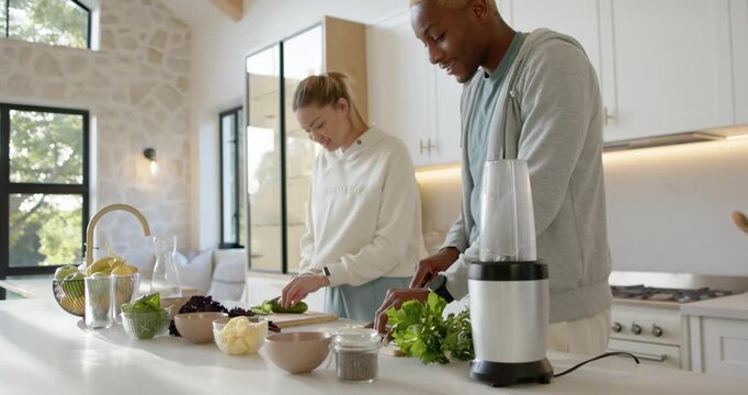 Diverse couple preparing meal at kitchen island, slicing cucumber, chopping herbs, blender idle